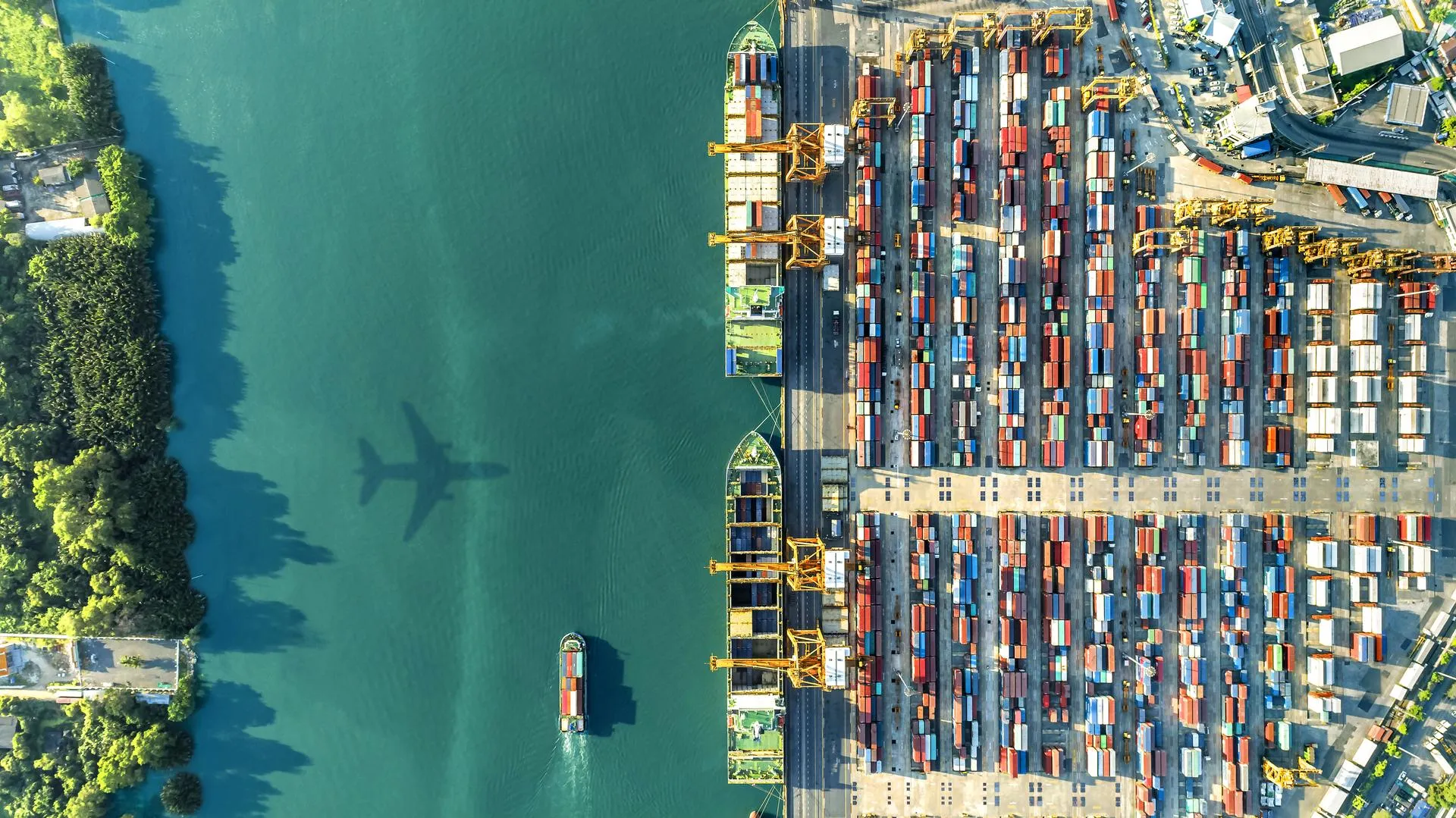 Aerial view of a river barge and a container yard with the shadow of a plane flying over the water.