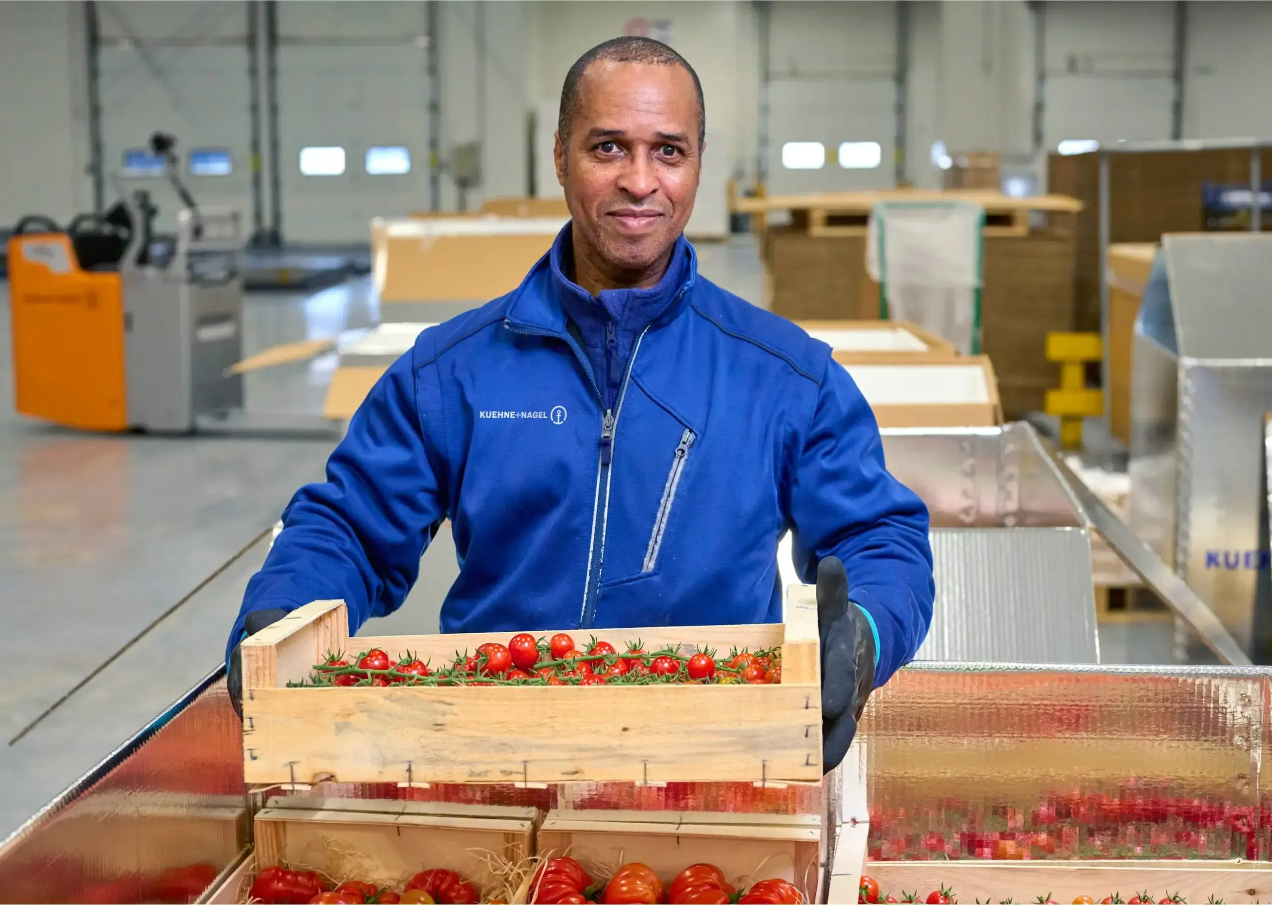 Man in Kuehne+Nagel suit carrying a box of tomatoes in a warehouse
