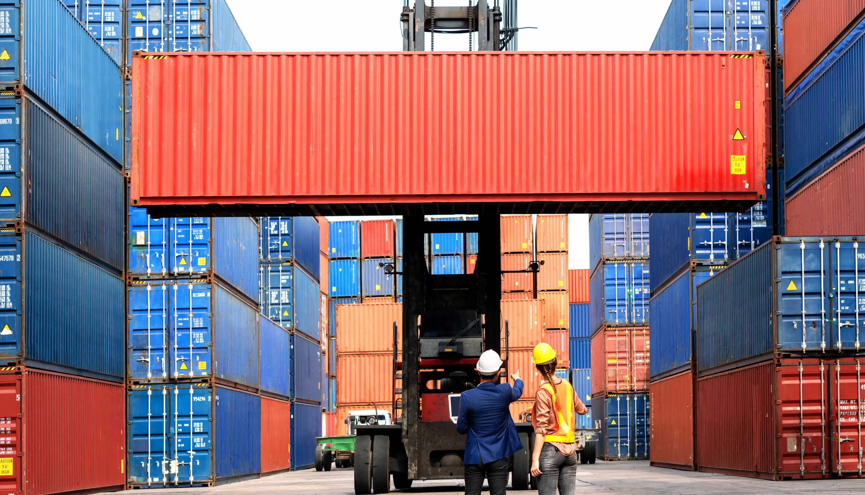 Two container yard workers are overseeing the transport of a red container.